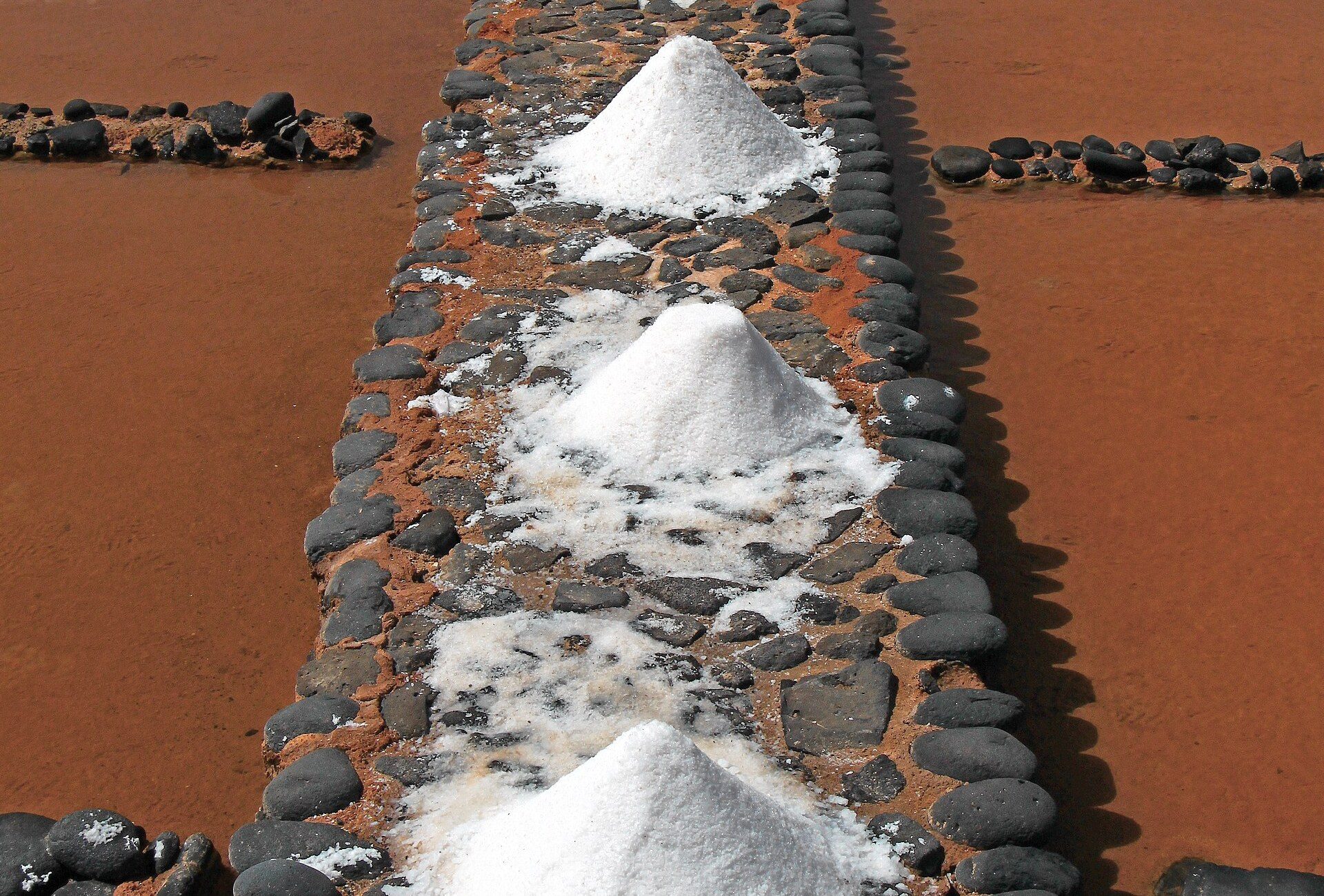 Rows of white salt piles on a stone wall between reddish salt evaporation pans